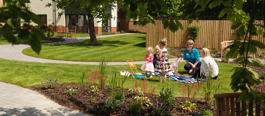 Young family with two small children sat on picnic blanket in the garden visiting resident