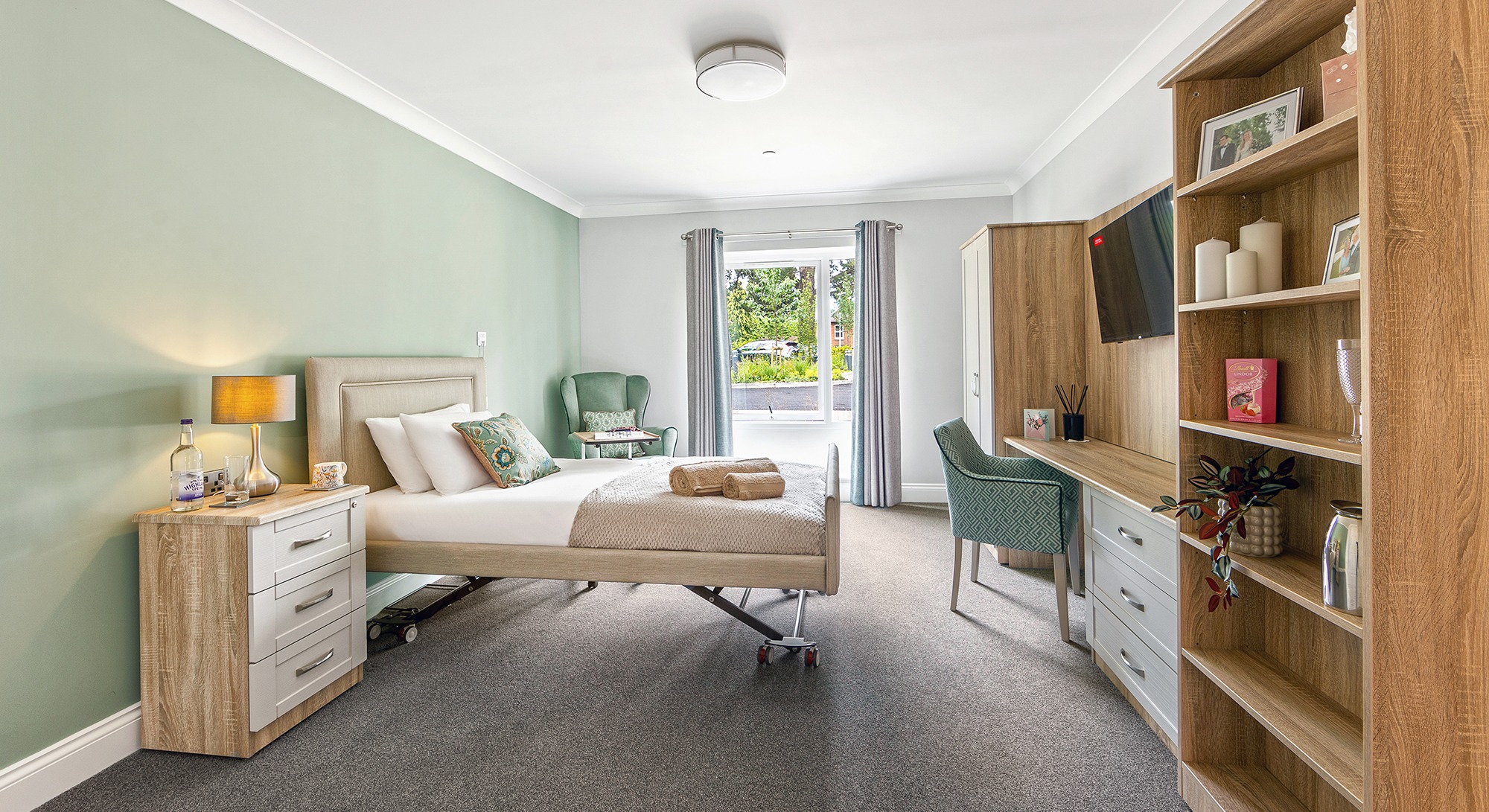 Resident’s bedroom at Moorhill Care Home featuring a single bed, a chair, fitted wardrobe with dressing table and shelves, and chest of drawers with lamp