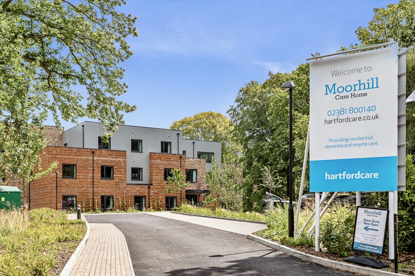 Front entrance to Moorhill Care Home with a sign displaying the name and ownership, and a small road leading to the main building surrounded by trees