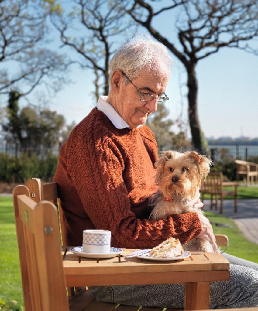 A resident sitting on a bench in the garden with a dog on his lap.